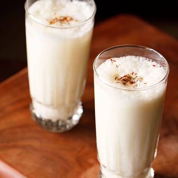 Two glasses of creamy Indian lassi, topped with saffron threads and spices, on a wooden surface.
