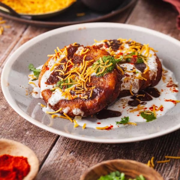 Two golden-brown Indian aloo tikki patties topped with white yogurt, dark chutney, green chutney, and sev.
