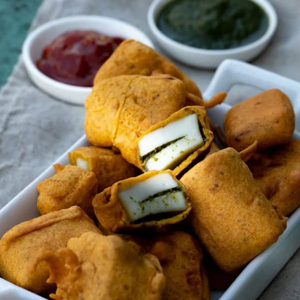 Crispy golden-brown paneer pakora, some cut open to reveal white cheese and green filling, with dipping sauces.