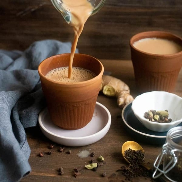 Masala chai being poured into a clay cup, with ginger and spices on a wooden table.